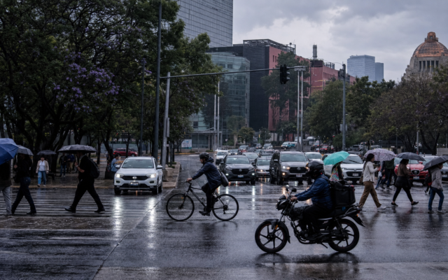 Automóviles avanzan lentamente sobre una avenida de la Ciudad de México mientras peatones cruzan con paraguas bajo un cielo nublado; al fondo se observan señales viales y edificios urbanos.
