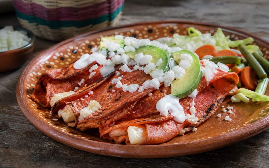 Enchiladas potosinas de masa rojiza con chile, queso, crema, aguacate y vegetales en plato de barro