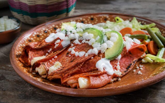 Enchiladas potosinas de masa rojiza con chile, queso, crema, aguacate y vegetales en plato de barro