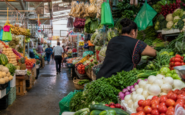 Pasillo de un mercado público de Ciudad de México con frutas, verduras y comerciantes atendiendo puestos