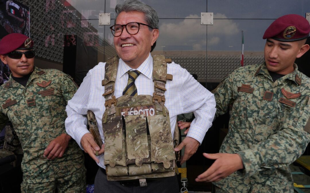 Ricardo Monreal prueba un chaleco táctico acompañado de dos militares durante una exhibición.