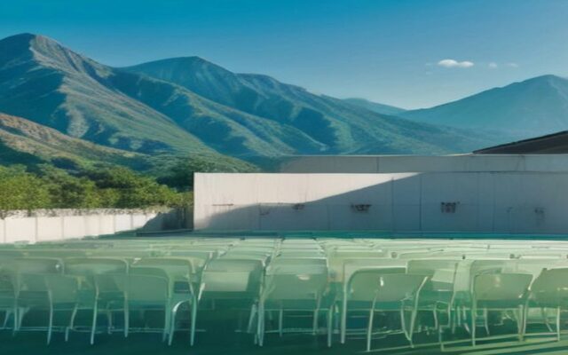 Cancha techada con estructura metálica y filas de sillas plásticas blancas vacías, con montañas verdes y cielo despejado al fondo