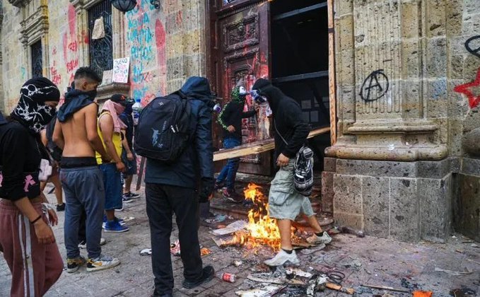 Jóvenes encapuchados rodean una fogata frente a una puerta de madera de Palacio de Gobierno con muros pintados en Guadalajara