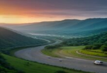Carretera sinuosa que atraviesa un valle verde entre montañas al atardecer, con un vehículo blanco circulando y nubes anaranjadas en el horizonte
