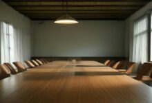 Larga mesa de juntas de madera con sillas vacías a los lados, ventanales y lámpara colgante en una sala iluminada