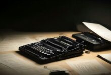 Máquina de escribir antigua de color negro sobre un escritorio de madera con hojas de papel dispersas, iluminada por una luz cálida y lateral en un ambiente oscuro