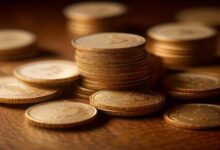 Pilas de monedas doradas sobre una mesa de madera oscura, con más monedas desenfocadas al fondo e iluminación cálida